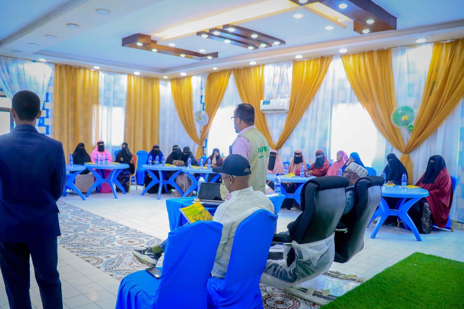 A trainer addressing workshop participants seated around tables in Adado